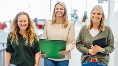 Drei Frauen lächeln in einem hellen Raum, eine hält einen grünen Ordner.