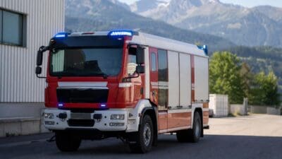 Rot-weißes Feuerwehrfahrzeug mit eingeschalteten blauen Blaulichtern vor Bergkulisse