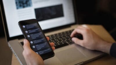 Eine Hand hält ein Smartphone mit Chat-Nachrichten vor einem Laptop, auf dem getippt wird
