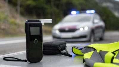 Ein digitales Alkoholmessgerät liegt neben einer gelben Warnweste vor einem Polizeiauto mit Blaulicht.