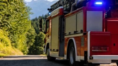 Seitenansicht eines roten Feuerwehrfahrzeugs mit Drehleiter auf einer Straße vor Bergen und Bäumen