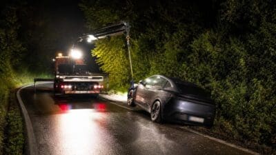 Ein schwerer Abschleppwagen mit beleuchtetem Kran steht neben einem abgeschleppten Elektroauto auf nasser Straße.