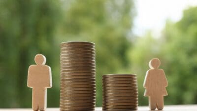 Linke Holzfigur eines Mannes neben hohem Münzstapel und rechte Holzfigur einer Frau neben niedrigerem Stapel