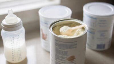 Eine Babyflasche mit Milch steht neben einer offenen Dose Babymilchpulver mit einem Messlöffel.