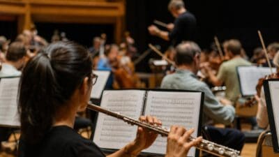 Eine Flötistin mit Brille und Pferdeschwanz spielt vor einem Notenpult in einem Orchester.