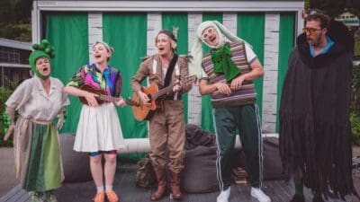 Fünf Schauspieler in bunten Kostümen singen und musizieren auf einer Bühne mit grünem Vorhang