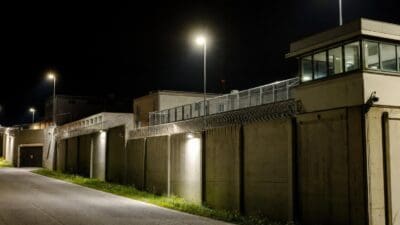 Nächtliche Ansicht einer Gefängnismauer mit Stacheldraht, Wachturm und Straßenbeleuchtung.