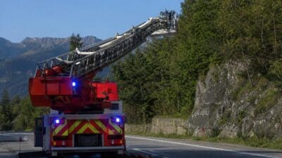 Rotes Feuerwehrfahrzeug mit ausgefahrener Drehleiter steht an einer Straße neben einem bewaldeten Hang.