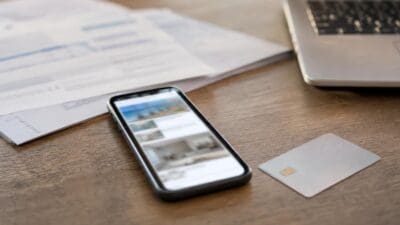 Smartphone zeigt App mit Ferienwohnungsfotos neben Kreditkarte und Papieren auf Holztisch