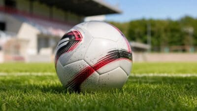 Ein weißer Fußball mit roten und schwarzen Akzenten liegt auf grünem Rasen vor einer Stadiontribüne.