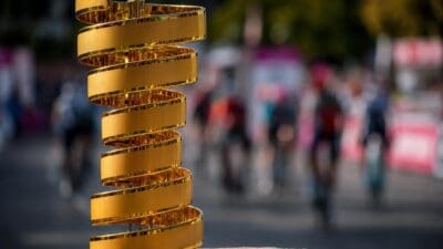 Goldener spiralförmiger Pokal auf einem Steinpodest mit unscharfen Radfahrern im Hintergrund