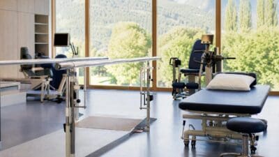 Therapieraum mit Behandlungsliege, Parallelbarren und Trainingsgeräten vor großen Fenstern mit Bergblick