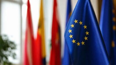 Blaue EU-Flagge mit gelben Sternen im Vordergrund und unscharfen Flaggen im Hintergrund.