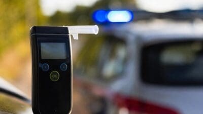 Ein Alkoholtestgerät mit Mundstück steht vor einem Polizeiwagen mit Blaulicht.