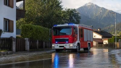 Rotes und weißes Feuerwehrfahrzeug mit Blaulicht auf nasser Straße zwischen Häusern und Bergen