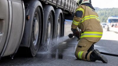 Feuerwehrmann in Schutzkleidung kniet auf der Autobahn und kühlt die Bremse eines Lkw mit Wasser.