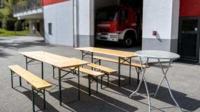 Mehrere Biertischgarnituren mit Bänken und ein runder Stehtisch vor einer roten Feuerwehrhalle mit Löschfahrzeug