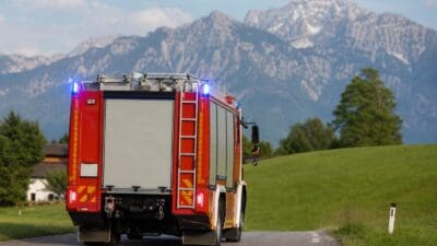 Heckansicht eines roten Feuerwehrfahrzeugs auf einer Landstraße mit schneebedeckten Bergen im Hintergrund