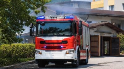 Rotes und weißes Feuerwehrfahrzeug mit leuchtenden blauen Blaulichtern vor einem Gebäude
