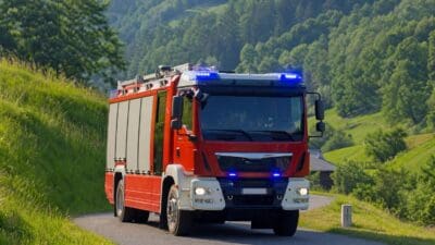 Rotes Feuerwehrfahrzeug mit aktiviertem Blaulicht auf einer Straße vor grünen Hügeln und Wäldern.