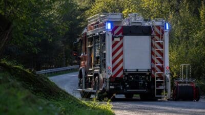 Heckansicht eines roten Feuerwehrfahrzeugs mit blauen Blinklichtern auf einer Waldstraße