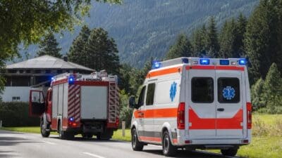 Rotes Feuerwehrfahrzeug und weiß-oranges Rettungsfahrzeug mit Blaulicht auf einer Landstraße vor Bergen.