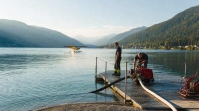 Zwei Feuerwehrleute bedienen eine rote Pumpe auf einem Steg am Weißensee mit einem Wasserflugzeug im Hintergrund