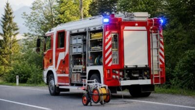 Rotes und weißes Feuerwehrfahrzeug mit geöffneten Geräteräumen und blauen Blinklichtern am Straßenrand