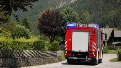 Rotes Feuerwehrfahrzeug von hinten mit blauen Blaulichtern auf einer Landstraße in bergiger Landschaft