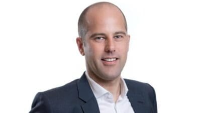 Lächelnder Mann mit kurzen Haaren in einem dunklen Anzug und weißem Hemd vor hellem Hintergrund
