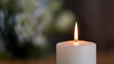 Eine brennende weiße Kerze mit heller Flamme vor unscharfem Blumenhintergrund