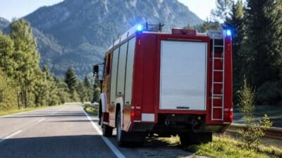 Heckansicht eines roten Feuerwehrfahrzeugs mit eingeschaltetem Blaulicht auf einer Landstraße vor Bergen
