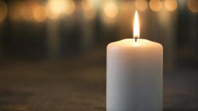 Eine weiße Kerze brennt auf einer dunklen Oberfläche, im Hintergrund sind unscharfe Lichter zu sehen.