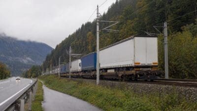 Langer Güterzug mit weißen und blauen Aufliegern auf Gleisen neben einer Autobahn im Bergtal