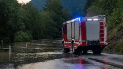 Rotes Feuerwehrfahrzeug mit Blaulicht auf überfluteter Straße in bergiger Waldlandschaft
