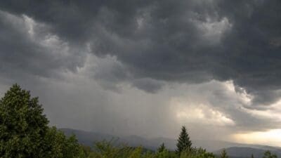 Dunkle Gewitterwolken mit sichtbarem Regen über einer grünen Berglandschaft und Bäumen im Vordergrund