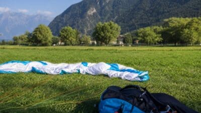 Weiß-blauer Gleitschirm liegt auf grüner Wiese mit Bergen und Bäumen im Hintergrund sowie Rucksack im Vordergrund