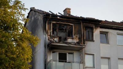 Stark beschädigte Fassade eines Mehrfamilienhauses mit zerstörten Fenstern und Dach nach einem Brand.