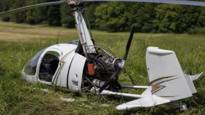 Ein weißer Gyrocopter liegt umgestürzt auf einer grünen Wiese mit freiliegendem Motor und Rotorblättern.