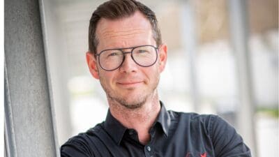 Ein Mann mit kurzen braunen Haaren, Brille und schwarzem Hemd lächelt in die Kamera.