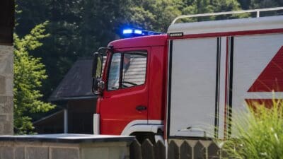 Rotes Feuerwehrfahrzeug mit blauem Blaulicht steht vor Gebäuden und Bäumen