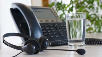 Ein schwarzes Headset liegt vor einem Telefon neben einem Glas Wasser.