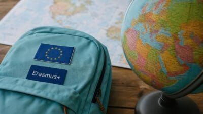 Türkiser Rucksack mit Erasmus+ Aufnäher und EU-Flagge neben einem Globus auf einer Weltkarte