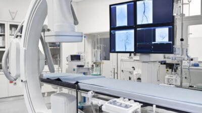 C-Bogen Angiographiegerät neben einem leeren Behandlungstisch und einer Monitorwand mit Gefäßbildern in einem hellen Interventionsraum.
