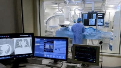 Arzt in blauer Schutzkleidung vor medizinischen Bildschirmen in einem Interventionsraum der Radiologie