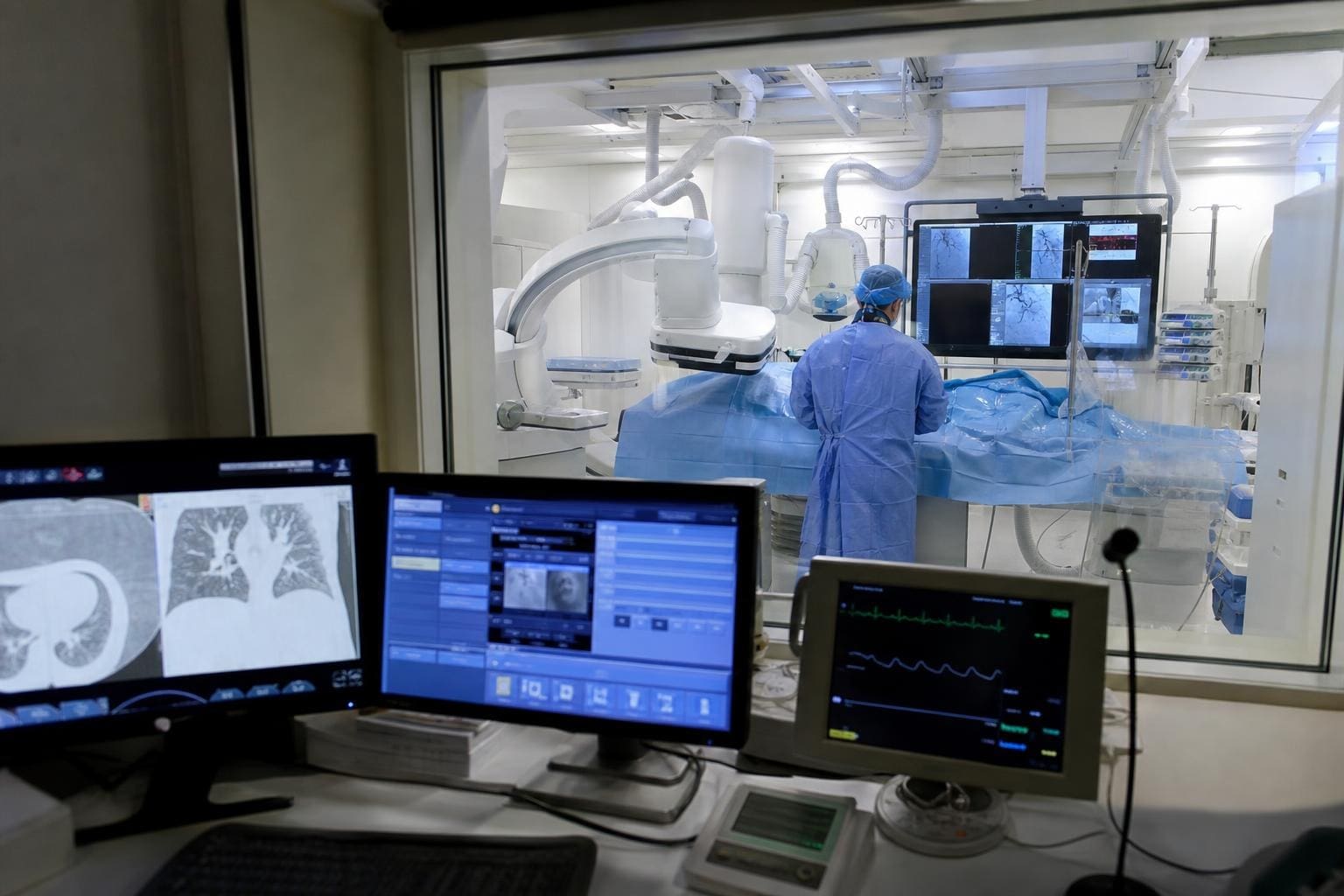 Arzt in blauer Schutzkleidung vor medizinischen Bildschirmen in einem Interventionsraum der Radiologie