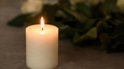 Eine brennende weiße Kerze vor unscharfen grünen Blättern und weißen Blumen