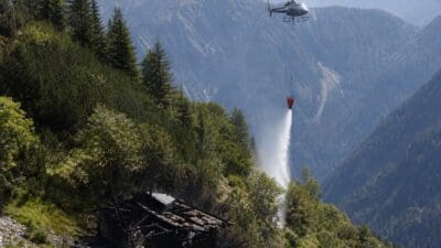 Hubschrauber gießt Wasser aus rotem Eimer auf Überreste einer abgebrannten Jagdhütte in den Bergen