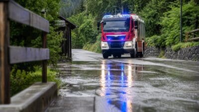 Rot-weißes Feuerwehrfahrzeug mit blauem Blaulicht fährt auf überfluteter Straße in bewaldetem Tal.