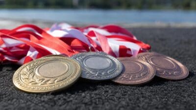 Goldene, silberne und bronzene Medaillen mit roten und weißen Bändern auf einer dunklen Oberfläche vor einem See.